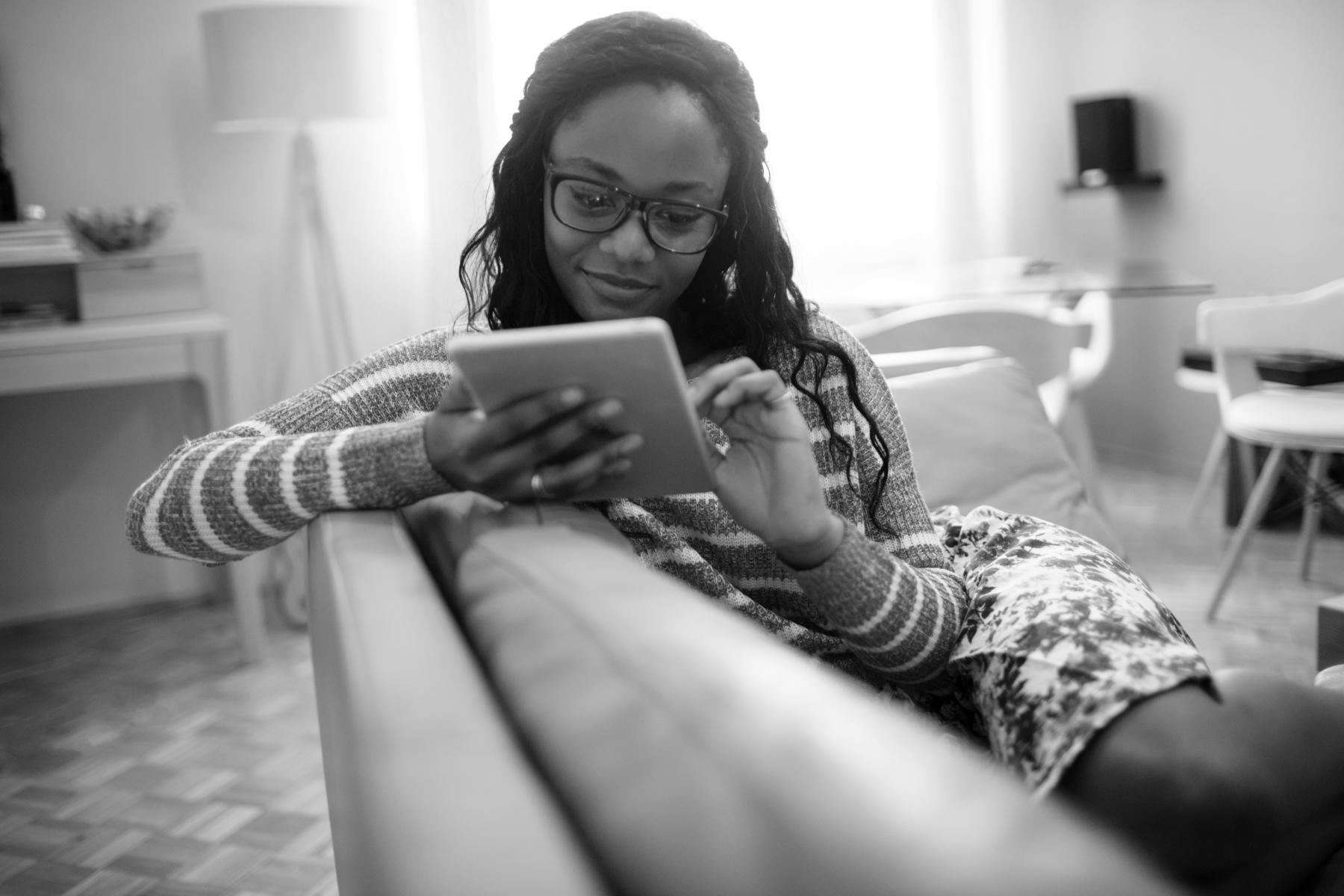 Photo of a young woman using her digital tablet while resting on the couch in her living room